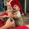baby, child, red_hat, smiling, video_game_controller, adult_arm, indoor, flooring, door, fan, toy, blurred_child, happy, casual_clothing, home, person, playing, candid, joyful, interaction