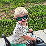 toddler, child, sunglasses, toy_vehicle, outdoor, grass, greenery, shorts, tshirt, blond_hair, summer, sunny, play, side_view, concrete, young_child, casual, reflection, hands_on_handlebars, cute