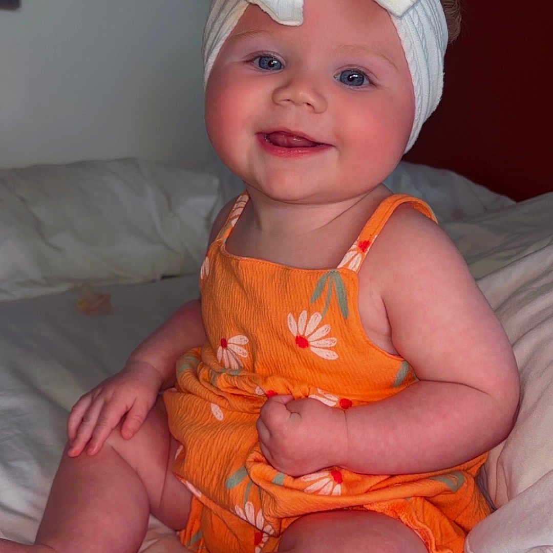 Journii is registered to the contest to win money with this photo: baby, bed, bow, child, cute, face, floral_pattern, foot, hand, happy, headband, indoors, infant, orange_clothing, person, portrait, sitting, skin, smiling, white_sheets