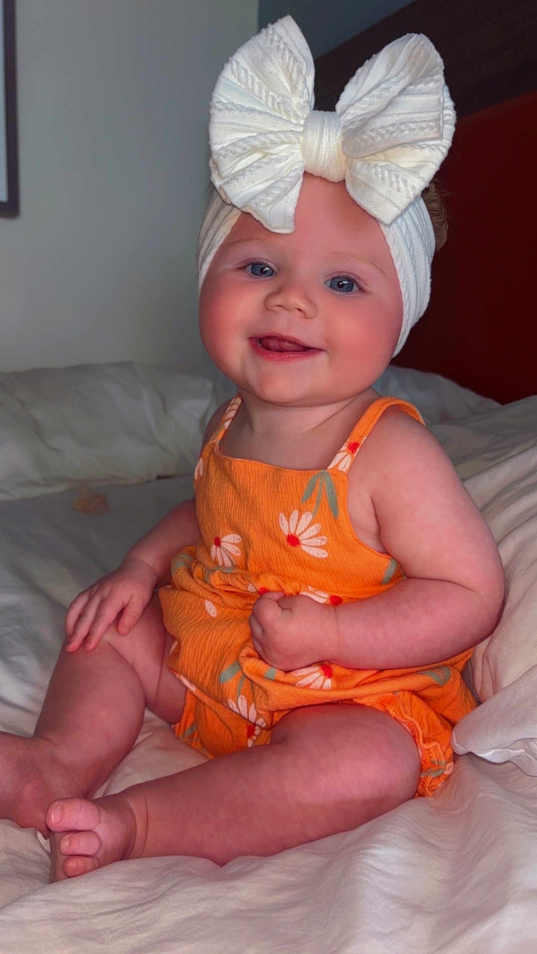 Journii is registered to the contest to win money with this photo: baby, child, smiling, headband, bow, orange_clothing, floral_pattern, sitting, bed, white_sheets, indoors, cute, happy, infant, portrait, skin, hand, foot, face, person