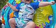 Jace is registered to the contest to win money with this photo: baby, infant, swimsuit, inflatable_pool, water_mat, grass, fan, colorful, playful, smiling, happy, outdoor, summer, legs, arms, patterned_clothing, toy, mat, barefoot, person