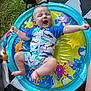baby, infant, swimsuit, inflatable_pool, water_mat, grass, fan, colorful, playful, smiling, happy, outdoor, summer, legs, arms, patterned_clothing, toy, mat, barefoot, person