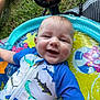 baby, smiling, child, inflatable_pool, swimsuit, shark_pattern, water, outdoor, grass, fan, summer, cute, happy, playtime, infant, blue_clothing, colorful, sunny, relaxed, fun
