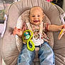 baby, child, smiling, happy, sitting, baby_seat, toy, colorful_toy, clothing, jeans, floral_top, indoor, person, cute, infant, face, hands, footwear, strap, comfort