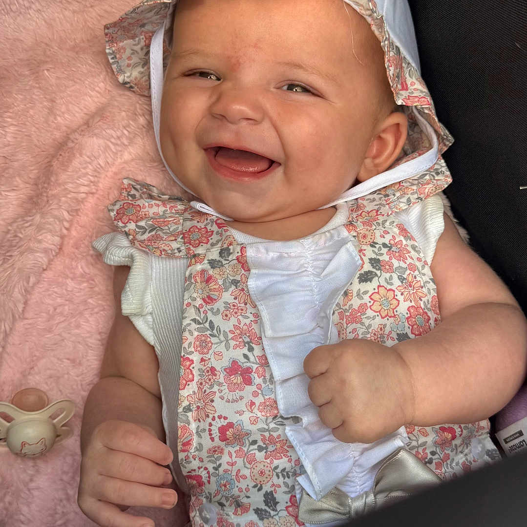 Harlow Rivah is registered to the contest to win money with this photo: adorable, baby, bonnet, child, closeup, cozy, cute, face, floral_outfit, hand, happy, indoors, infant, pacifier, pink_blanket, portrait, sitting, smiling, soft_texture, white_clothing