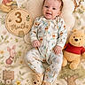 baby, infant, smiling, winnie_the_pooh, plush_toy, pajamas, blanket, wooden_sign, flowers, soft_lighting, cute, child, lying_down, decor, illustration, stuffed_animal, happy, face, footwear, cozy