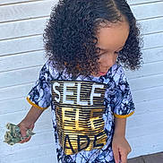 Messiah Harris is registered to the contest to win money with this photo: black_and_white, casual_clothing, child, curly_hair, daylight, fashion, gold_lettering, hand, looking_down, money, outdoor, person, portrait, short_sleeves, standing, text_on_clothing, tshirt, wooden_floor, wooden_wall, young_person