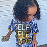 black_and_white, casual_clothing, child, curly_hair, daylight, fashion, gold_lettering, hand, looking_down, money, outdoor, person, portrait, short_sleeves, standing, text_on_clothing, tshirt, wooden_floor, wooden_wall, young_person