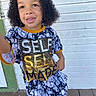casual_clothing, child, closeup, curly_hair, daylight, fashion, green_door_frame, happy, outdoor, person, photography, portrait, self_made, shirt, smiling, standing, text_on_clothing, toddler, wooden_floor, wooden_wall