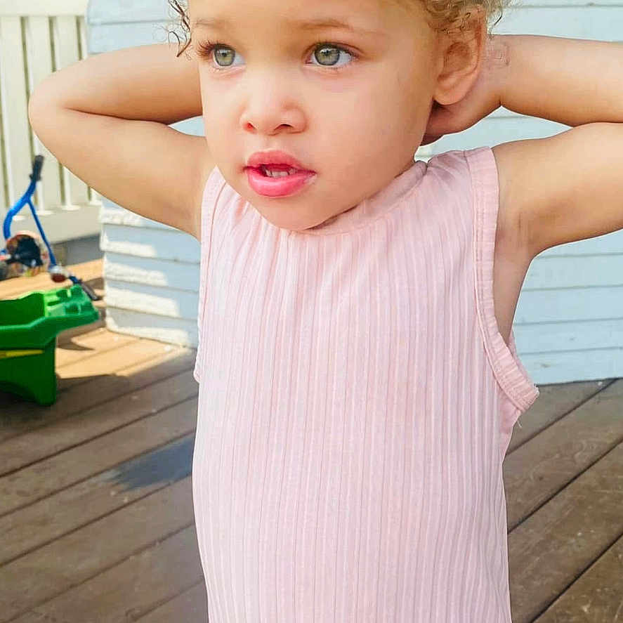 Zahra Harris joined the competition — help win amazing prizes! child, girl, outdoor, dress, pink, curly_hair, deck, wooden_floor, porch, sunlight, standing, person, casual_clothing, hairstyle, young_child, expression, daylight, white_wall, fence, background