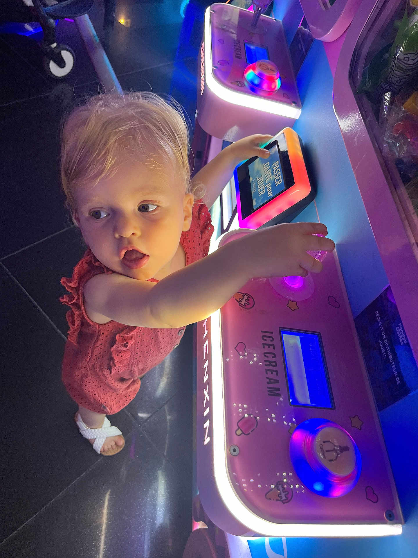 Léna participe au concours pour gagner de l'argent avec cette photo : toddler, child, arcade_machine, pink_dress, white_sandals, neon_lights, indoor, game, curious, playful, technology, floor_tiles, hand, face, young_child, machine, colorful, entertainment, person, light
