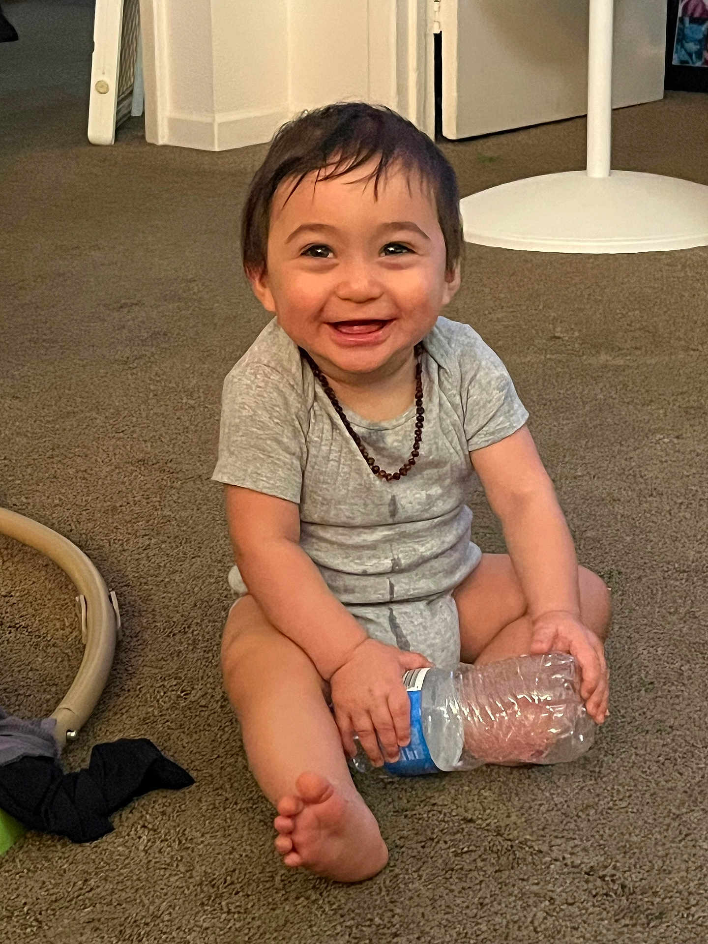 David joined the competition — help win amazing prizes! toddler, child, smiling, sitting, carpet, water_bottle, onesie, necklace, indoor, barefoot, happy, cute, baby, person, floor, home, casual, playful, young, portrait