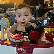 David is registered to the contest to win money with this photo: baby, child, walker, toy, disney, mickey_mouse, indoor, curious, face, furniture, blanket, couch, home, floor, playtime, infant, person, cute, decorations, colorful