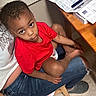 toddler, child, red_shirt, sitting, lap, adult, jeans, indoor, table, packaging, floor, curious, looking_up, wooden_table, casual, person, footwear, instruction_manual, home, cozy