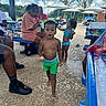 child, toddler, swimwear, outdoor, summer, picnic, poolside, bench, adult, umbrella, sky, cloud, concrete, footwear, green_shorts, blue_swimsuit, people, sunlight, table, recreation