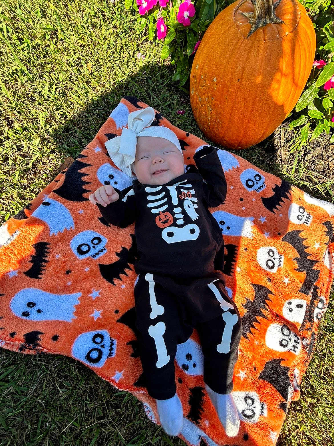 Lilly is registered to the contest to win money with this photo: baby, infant, halloween, costume, skeleton_outfit, pumpkin, blanket, ghost, bat, skull, grass, flower, pink_flower, headband, smile, outdoor, sunlight, holiday, cute, festive