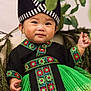 toddler, child, traditional_clothing, hat, green_dress, embroidery, floral_pattern, smiling, portrait, face, hand, barefoot, leaves, branches, nature, indoor, cute, young_child, happy, costume