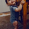 toddler, child, standing, smiling, hair_buns, wooden_chair, indoor, carpet, baby_clothing, happy, person, young_child, cute, holding, furniture, floor, home, portrait, playful, small