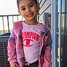 child, girl, smile, pink_clothing, jacket, tshirt, plaid, balcony, railing, outdoor, daylight, building, window, hair, face, person, casual, young, standing, happy