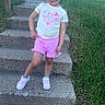 child, girl, smiling, posing, steps, concrete, grass, outdoor, shorts, tshirt, footwear, hair_tied, house, window, sidewalk, playful, young, casual_clothing, daylight, standing