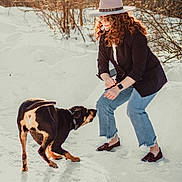 Ducky is registered to the contest to win money with this photo: woman, dog, snow, winter, outdoor, hat, sunglasses, jeans, black_jacket, playful, pet, animal, nature, daylight, smile, curly_hair, footwear, trees, interaction, happy