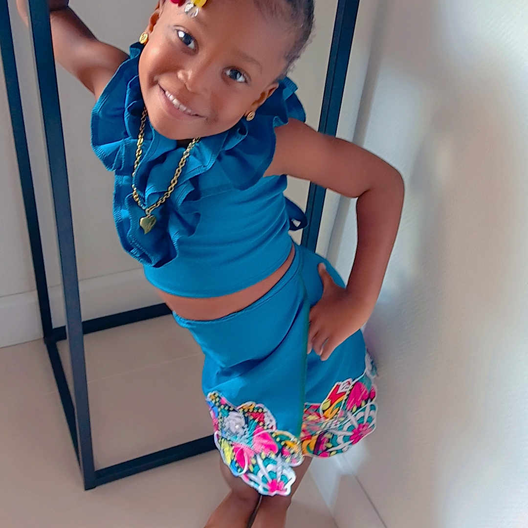 Melyanna a rejoint le concours — aidez-le/la à gagner de superbes lots ! barefoot, blue_outfit, braids, child, colorful_beads, cute, earrings, fashion, floor, girl, happy, indoor, jewelry, necklace, playful, portrait, pose, smile, table, wall