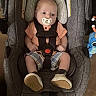 Kyle joined the competition — help win amazing prizes! baby, car_seat, pacifier, infant, child, plaid_shorts, peach_shirt, shoes, straps, sitting, indoor, toy, plush_toy, gray_fabric, cute, wide_eyes, comfort, safety, car_safety, child_seat