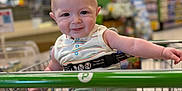 Cameron is registered to the contest to win money with this photo: baby, child, shopping_cart, grocery_store, smile, striped_shirt, green_shorts, seatbelt, shopping, aisle, store, cute, happy, person, infant, retail, playful, tongue_out, indoor, young_child
