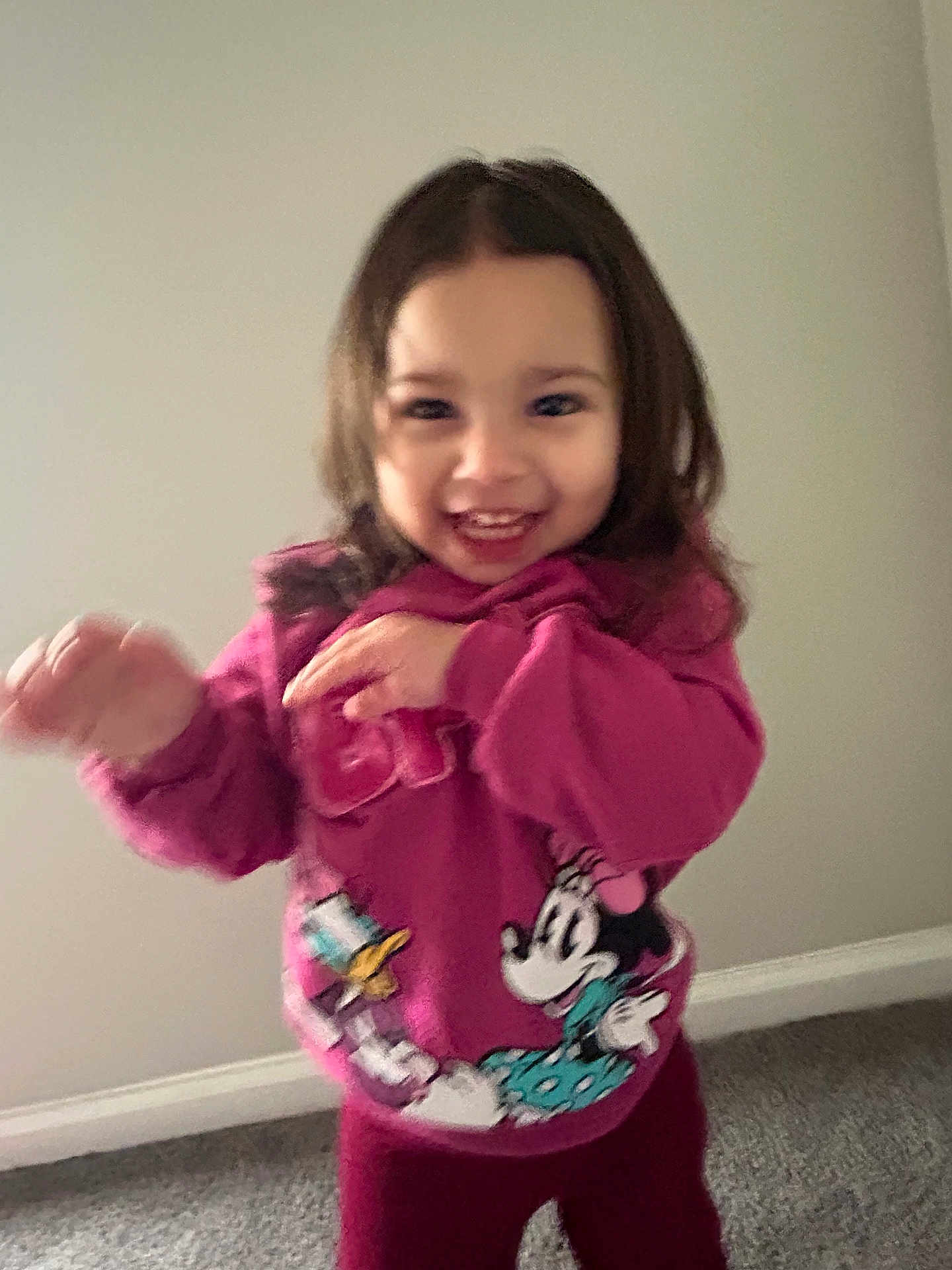 Isabella is registered to the contest to win money with this photo: toddler, child, smiling, pink_sweatshirt, indoor, carpet, wall, cartoon_characters, motion_blur, happy, person, clothing, long_hair, playful, cute, young_child, standing, casual_wear, expression, portrait
