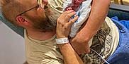 Colton is registered to the contest to win money with this photo: newborn, baby, man, adult, beard, glasses, hospital, blanket, chair, indoor, person, holding, seated, love, family, portrait, tenderness, casual_clothing, wood_floor, table