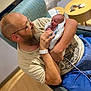 newborn, baby, man, adult, beard, glasses, hospital, blanket, chair, indoor, person, holding, seated, love, family, portrait, tenderness, casual_clothing, wood_floor, table