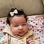 baby, child, hoodie, bow, blanket, heart_pattern, chubby_cheeks, indoor, cute, soft_lighting, hand, face, person, infant, clothing, pink, red, beige, portrait, relaxed