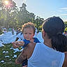 adult, being_held, blue_sky, bubble_party, child, crowd, curious, family, foam, grass, outdoor, parent, park, portrait, reaching, summer, sunlight, tattoo, toddler, trees