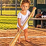 child, toddler, baseball, baseball_bat, baseball_field, sports, athlete, outdoor, sunlight, golden_hour, barefoot, white_shirt, standing, portrait, face, serious_expression, baseball_glove, baseballs, fence, grass