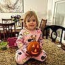 cartoon_clothing, chairs, child, countertop, dining_room, dining_table, family_home, flowers, girl, halloween, high_chair, indoor, jack_o_lantern, kitchen, pajamas, pink, portrait, pumpkin, smiling, toddler
