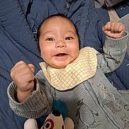 Legend is registered to the contest to win money with this photo: baby, infant, smiling, happy, fist, onesie, bib, penguin_print, blanket, plush_toy, bed, cozy, cute, child, face, portrait, indoor, clothing, expression, lying_down