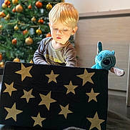 Aëden participe au concours pour gagner de l'argent avec cette photo : blanket, blonde_hair, blue_toy, box, child, christmas_tree, cozy, curious, decorations, festive, floor, holiday, holiday_season, indoor, ornaments, plush_toy, soft_toy, stars, toddler, young_child