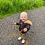 toddler, child, smiling, wildflowers, gravel_path, black_jacket, hood, outdoor, greenery, grass, nature, shoes, candid, happy, cute, person, young_child, playful, daylight, casual_clothing