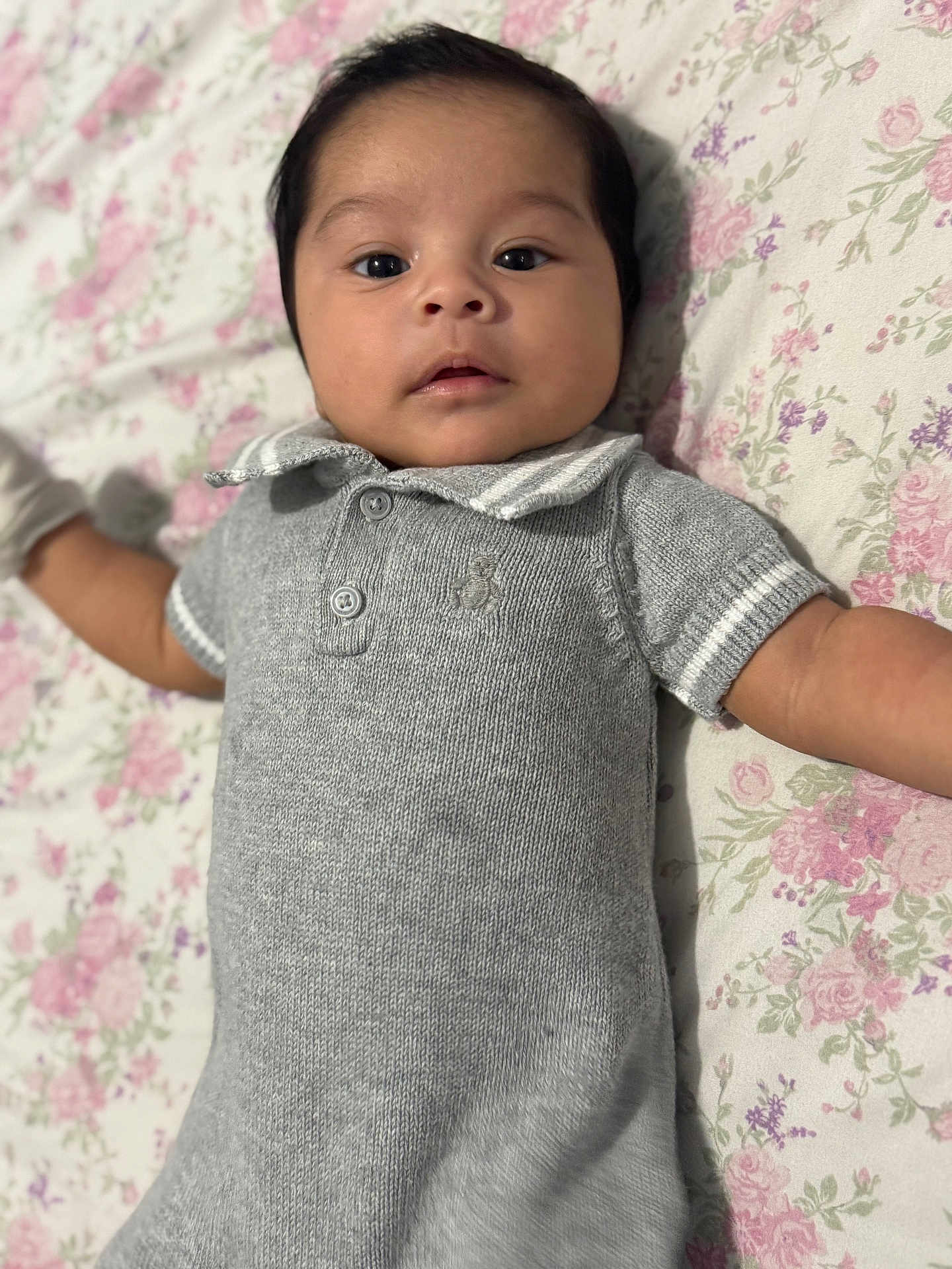 Carlos is registered to the contest to win money with this photo: baby, infant, child, face, portrait, lying_down, blanket, floral_pattern, knitwear, grey_clothing, cute, young, looking, eyes, collar, buttons, soft_texture, indoors, skin, person