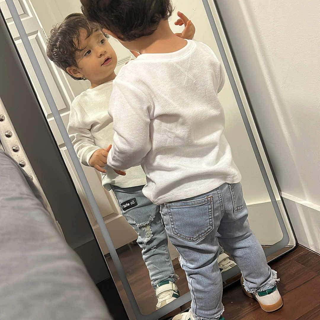 Eren is registered to the contest to win money with this photo: casual_clothing, child, curiosity, curly_hair, floor, full_length_mirror, home, indoor, looking, mirror, person, reflection, ripped_jeans, room, sneakers, standing, toddler, white_sweater, wooden_floor, young_child