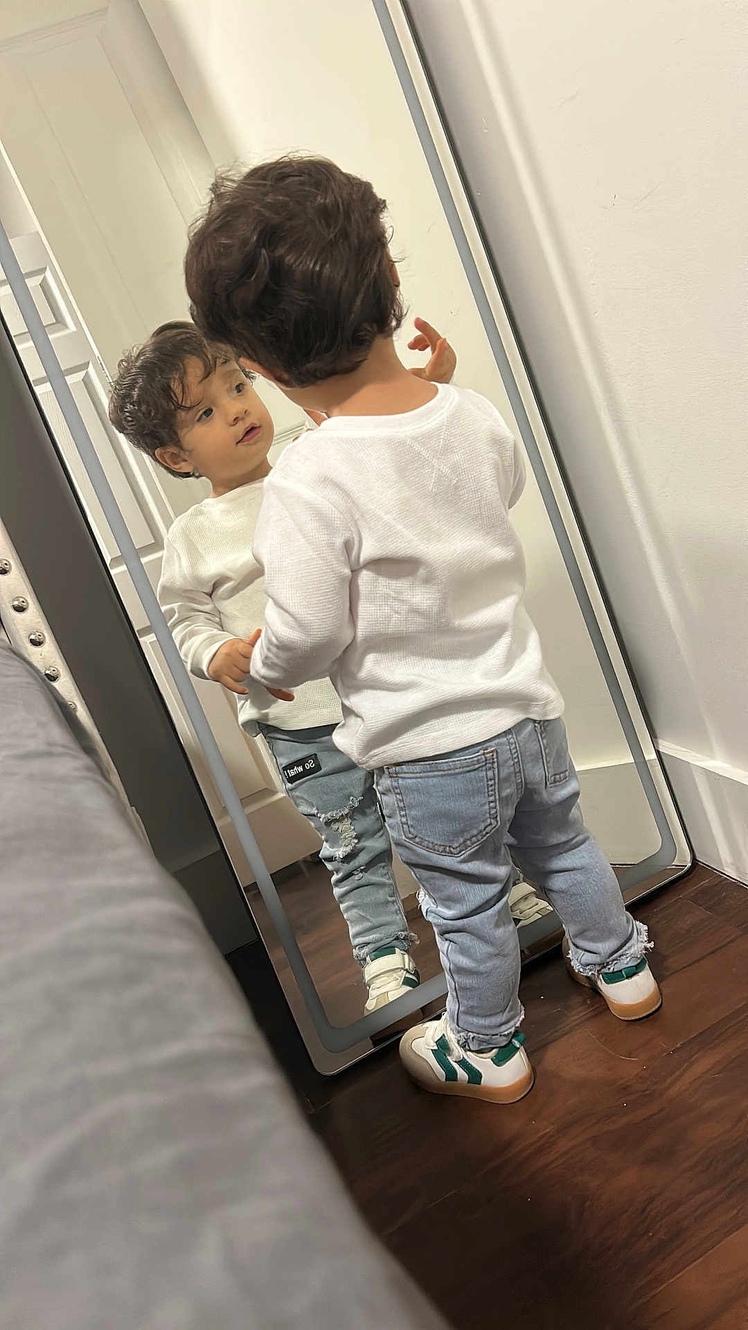 Eren is registered to the contest to win money with this photo: casual_clothing, child, curiosity, curly_hair, floor, full_length_mirror, home, indoor, looking, mirror, person, reflection, ripped_jeans, room, sneakers, standing, toddler, white_sweater, wooden_floor, young_child