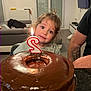 child, cake, birthday_candle, chocolate_cake, indoor, celebration, toddler, person, happy, smile, hair_tie, table, living_room, sofa, television, tattoo, arm, glass_plate, light, curious