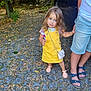 child, girl, yellow_dress, pink_sandals, cobblestone_path, dry_leaves, person, legs, denim_shorts, sandals, outdoor, daylight, tree, car, handbag, curly_hair, blue_eyes, holding_hand, nature, casual_clothing