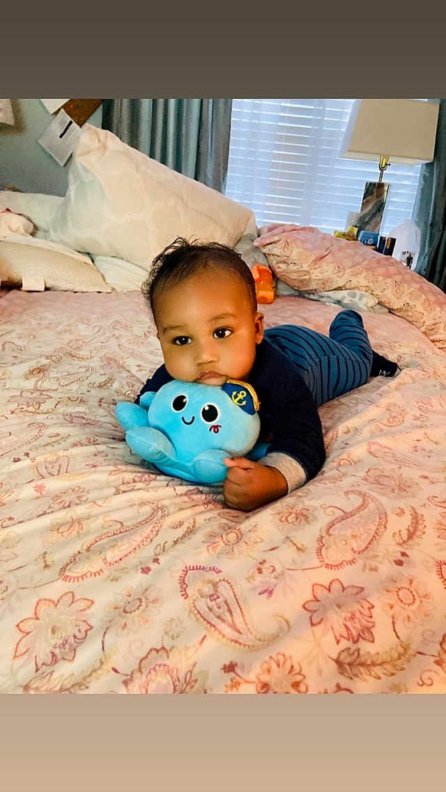Roderick is registered to the contest to win money with this photo: baby, infant, child, plush_toy, octopus_toy, bed, bedding, pillow, blanket, indoor, portrait, lying_down, crawling, smiling, onesie, cute, lamp, window, curtains, nursery