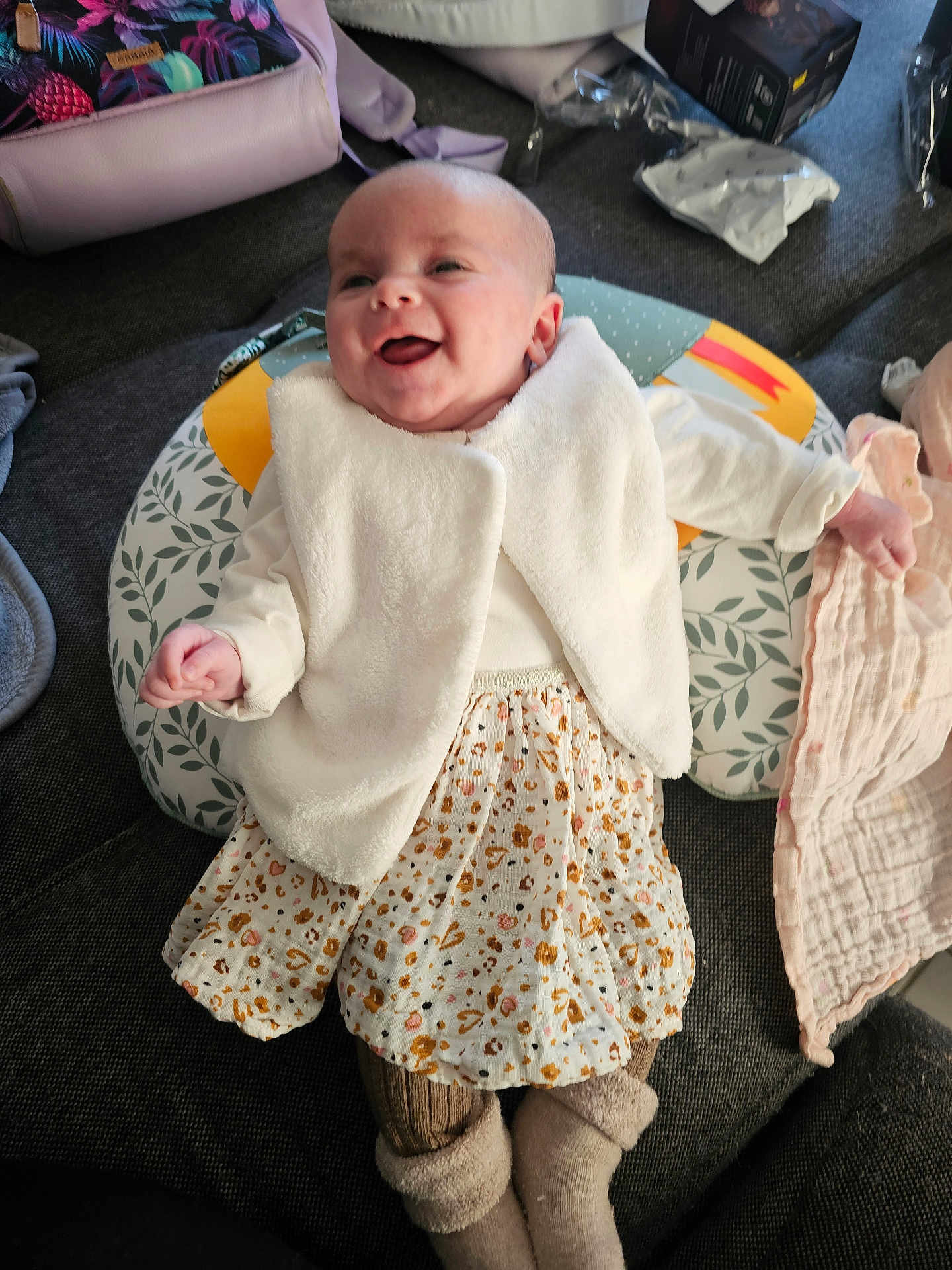 Lucie a rejoint le concours — aidez-le/la à gagner de superbes lots ! baby, infant, smiling, laughing, white_jacket, floral_skirt, socks, cushion, nursing_pillow, couch, blanket, backpack, packaging, plastic_wrap, box, happy, portrait, home_interior, person, clothing