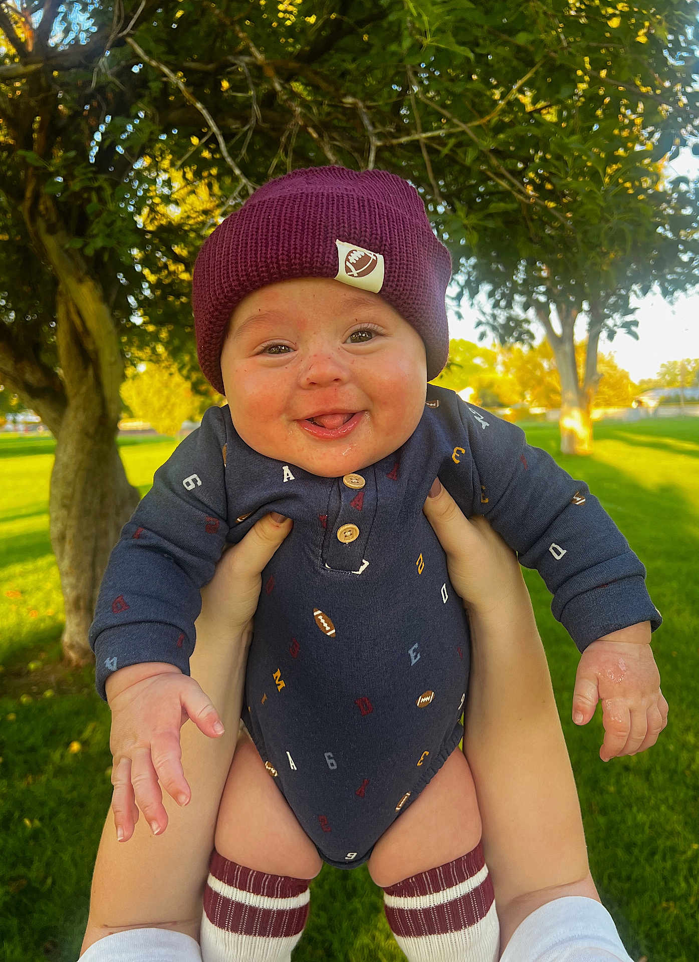 Callahan joined the competition — help win amazing prizes! baby, child, smiling, outdoor, greenery, trees, grass, knit_hat, onesie, football_pattern, hands, person, happy, sunlight, nature, park, cute, playful, warm_light, portrait