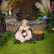 Valerie joined the competition — help win amazing prizes! toddler, child, lantern, pink_dress, flower_crown, green_rug, artificial_flowers, foliage, garden_scene, decorative_animals, rabbit_figurine, hedgehog_figurine, tree_stump, bench, portrait, indoor, whimsical, fairy_tale, cute, sitting
