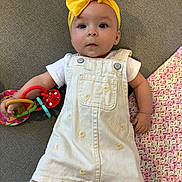 Camila is registered to the contest to win money with this photo: baby, child, yellow_bow, headband, white_dress, overalls, toy, colorful_rings, crocheted_blanket, gray_surface, infant, cute, portrait, indoor, small_hand, clothing, expression, lying_down, person, playtime