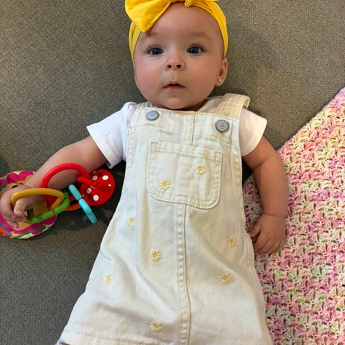 Camila is registered to the contest to win money with this photo: baby, child, yellow_bow, headband, white_dress, overalls, toy, colorful_rings, crocheted_blanket, gray_surface, infant, cute, portrait, indoor, small_hand, clothing, expression, lying_down, person, playtime