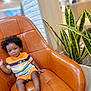 toddler, child, curly_hair, striped_clothing, orange_outfit, gold_necklace, gold_bracelet, leather_chair, indoor, plant, green_plant, modern_interior, white_wall, wood_panel, barefoot, smiling, sitting, cute, portrait, relaxed